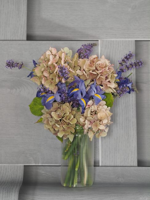 A floral arrangement featuring a bouquet of hydrangeas in soft pink and beige tones, complemented by vibrant purple irises and sprigs of lavender with purple flower clusters. The flowers are arranged