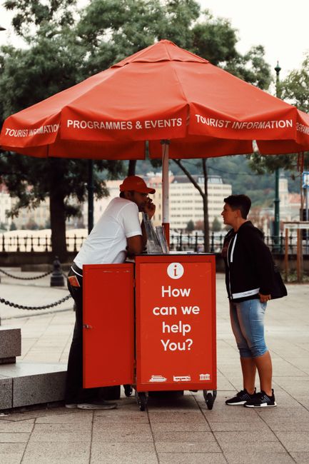 This image depicts a tourist information kiosk situated outdoors under a large, orange umbrella that features printed text advertising 'PROGRAMMES & EVENTS' and 'TOURIST INFORMATION.' The kiosk is man