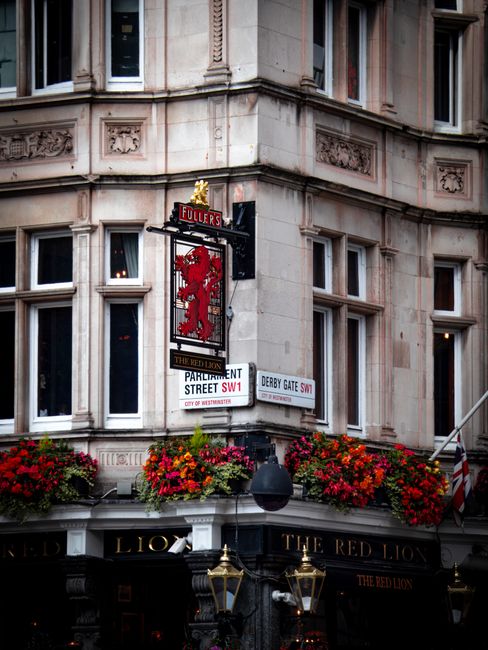 Photograph of the corner of a historic building at the intersection of Parliament Street and Derby Gate SW1 in Westminster, featuring ornate stonework and multiple windows. A prominent hanging sign di