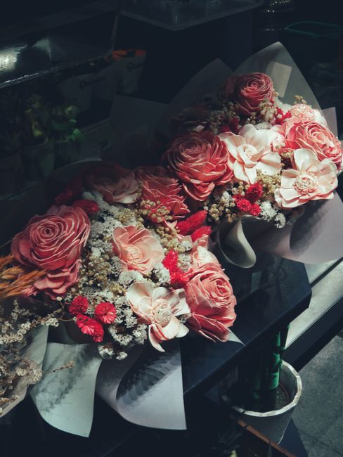 A floral arrangement featuring a bouquet of fresh pink roses, cream-colored orchids, and small red and white filler flowers, artistically wrapped in white paper with a satin ribbon. The bouquet is dis