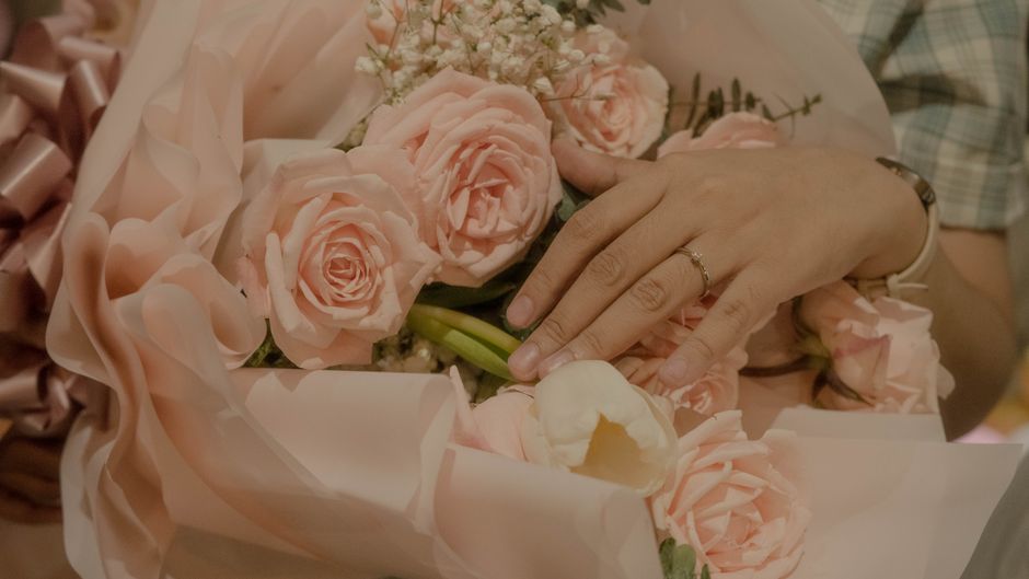 A close-up of a person's hand gently resting on a bouquet of fresh, pastel pink roses with soft, layered petals and green stems, accented with delicate white baby's breath. The bouquet is wrapped in l