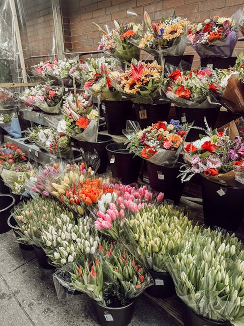 A display of numerous flower bouquets and bunches arranged in black buckets and on shelves, featuring a variety of fresh flowers such as tulips, lilies, roses, and mixed seasonal blooms. The flowers s